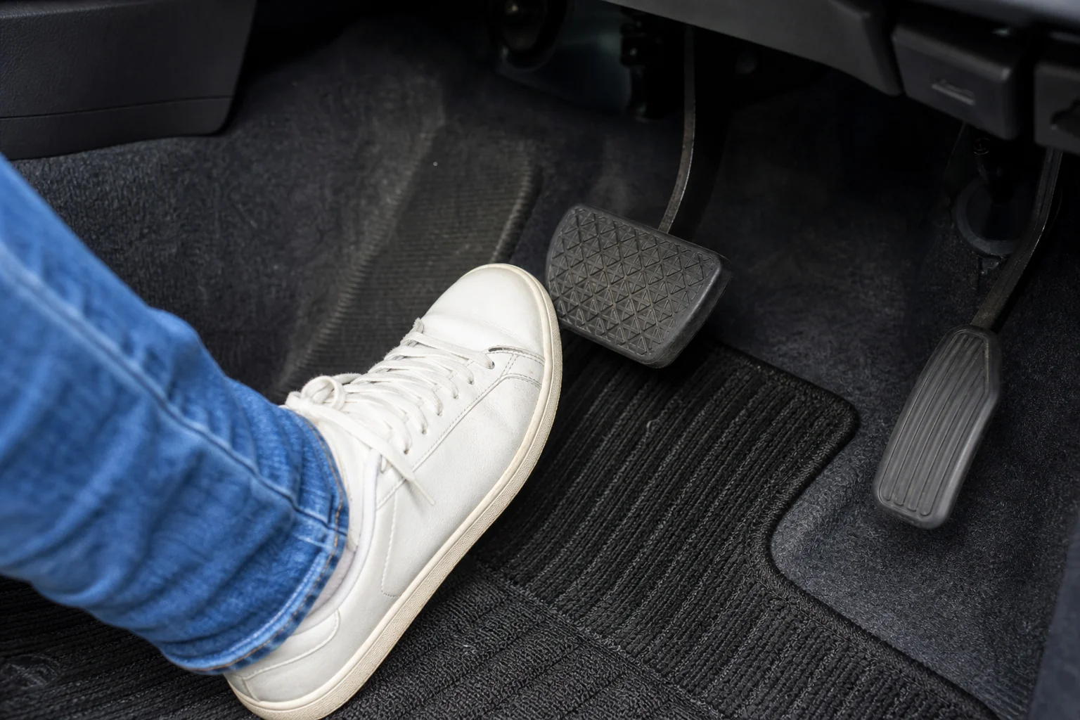 教習車の運転席の足元で、平らなスニーカーを履いた足がブレーキペダルの前に置かれている写真